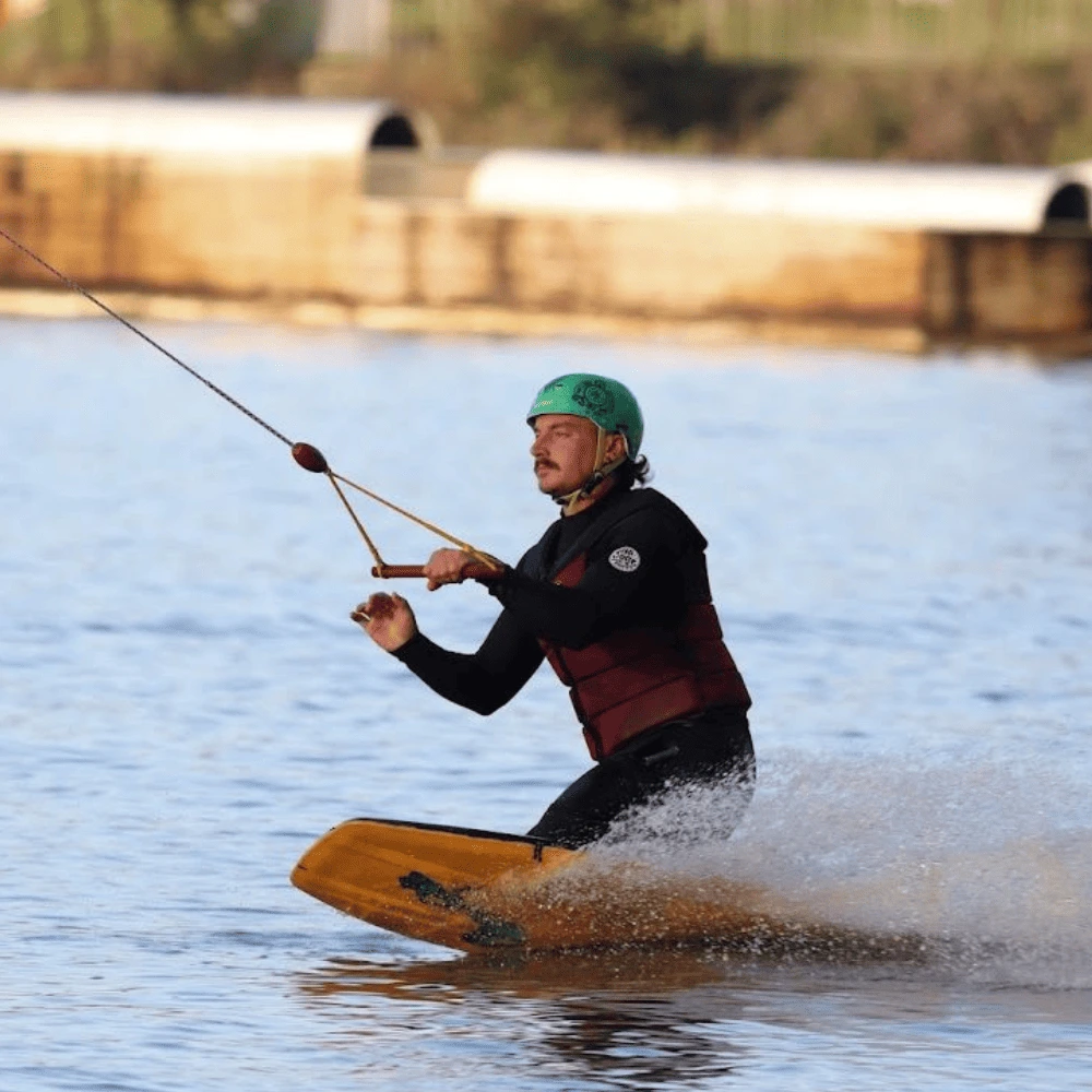 Pratique du wakeboard au wakepark de la Rincerie en Mayenne. Un rider équipé d’un casque vert glisse sur l’eau en plein contrôle avec un câble.