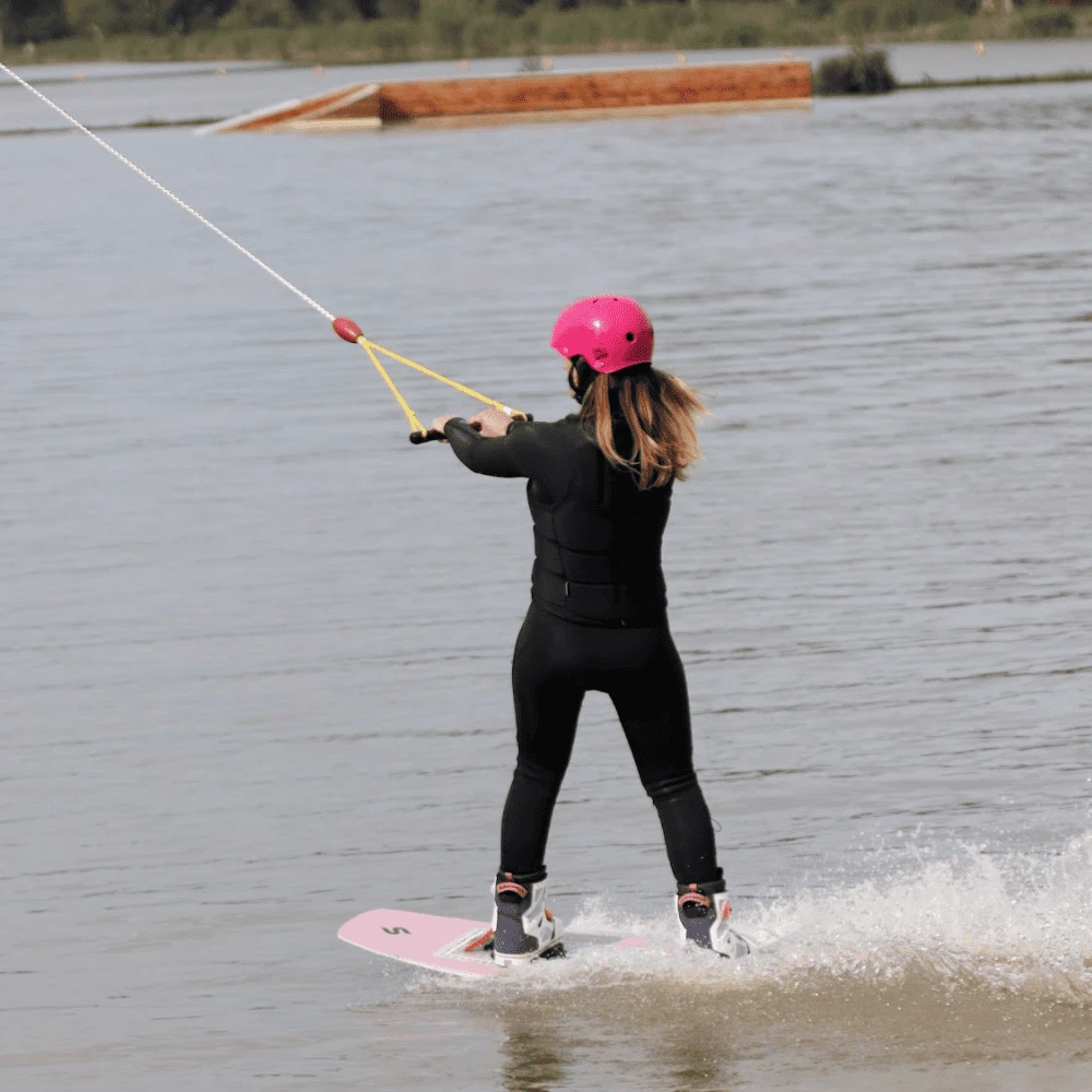 Femme pratiquant le wakeboard au wakepark de la Rincerie en Mayenne. Équipée d’un casque rose, elle glisse élégamment sur l’eau grâce au système de câble.